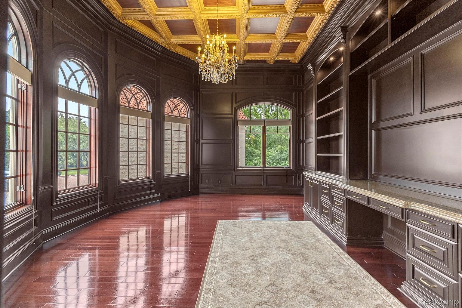 This is an interior shot of a luxurious office or study, featuring dark wood paneling, a coffered ceiling with gold accents, and a crystal chandelier. The room has large arched windows providing natural light, a built-in bookcase with a desk area, and a decorative area rug on the hardwood floor. The overall impression is one of sophistication and elegance.