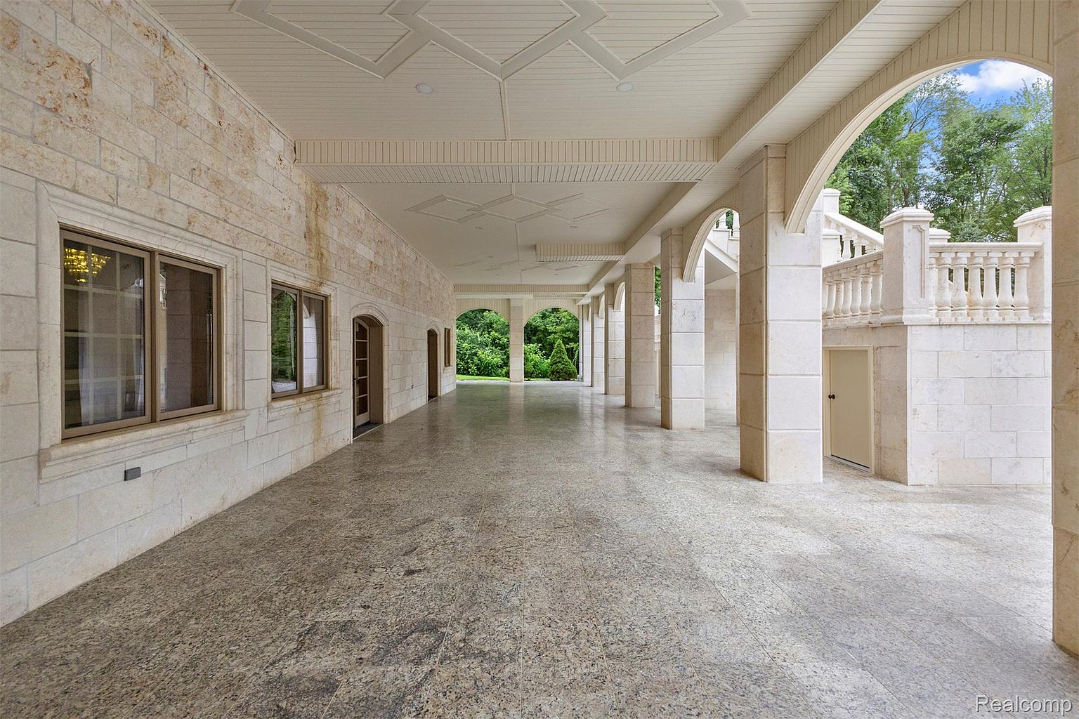 This image showcases a spacious outdoor patio or covered walkway, constructed with light-colored stone and featuring a series of columns and arches. The flooring is a speckled stone, and the ceiling is adorned with a geometric pattern. The area provides a transition between the interior and the lush greenery visible in the background, suggesting a luxurious and well-designed outdoor living space.