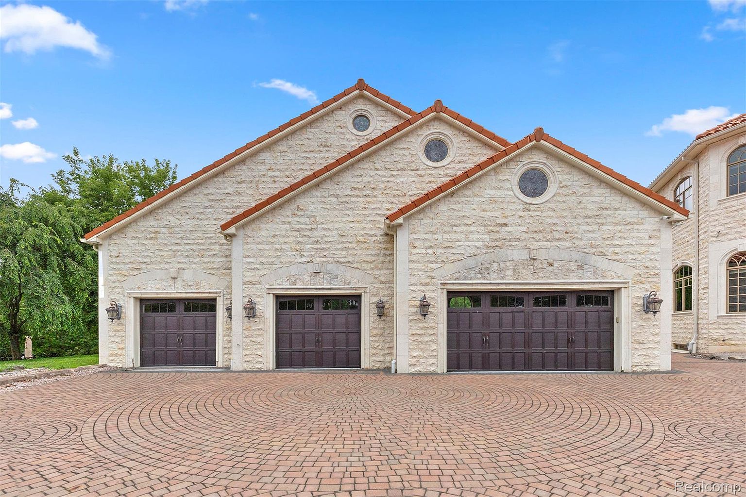This image showcases a luxurious three-car garage with a stone facade and brown garage doors. The driveway is paved with a circular brick pattern, adding to the property's curb appeal. The architectural style is elegant and well-maintained, suggesting a high-end property.