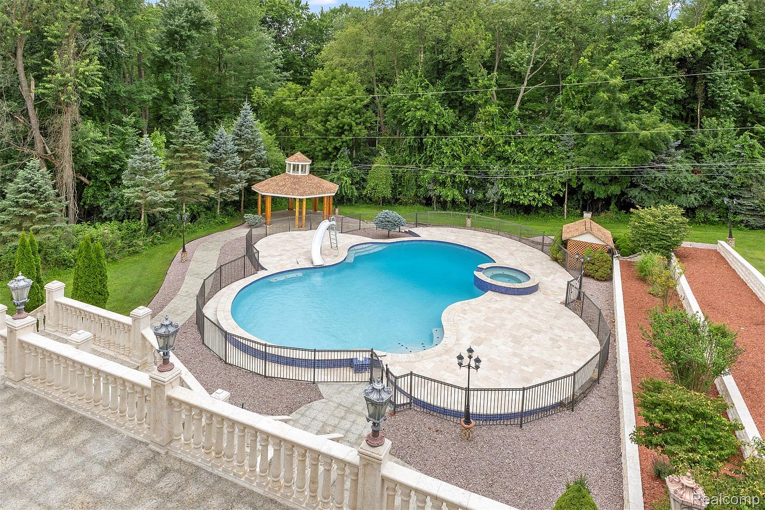 This is an aerial view of a luxurious backyard featuring a uniquely shaped pool with an attached spa and a slide. The pool area is surrounded by a stone patio, a black metal fence, and meticulously maintained landscaping, including a gazebo. The property is bordered by lush, mature trees, creating a private and serene setting.