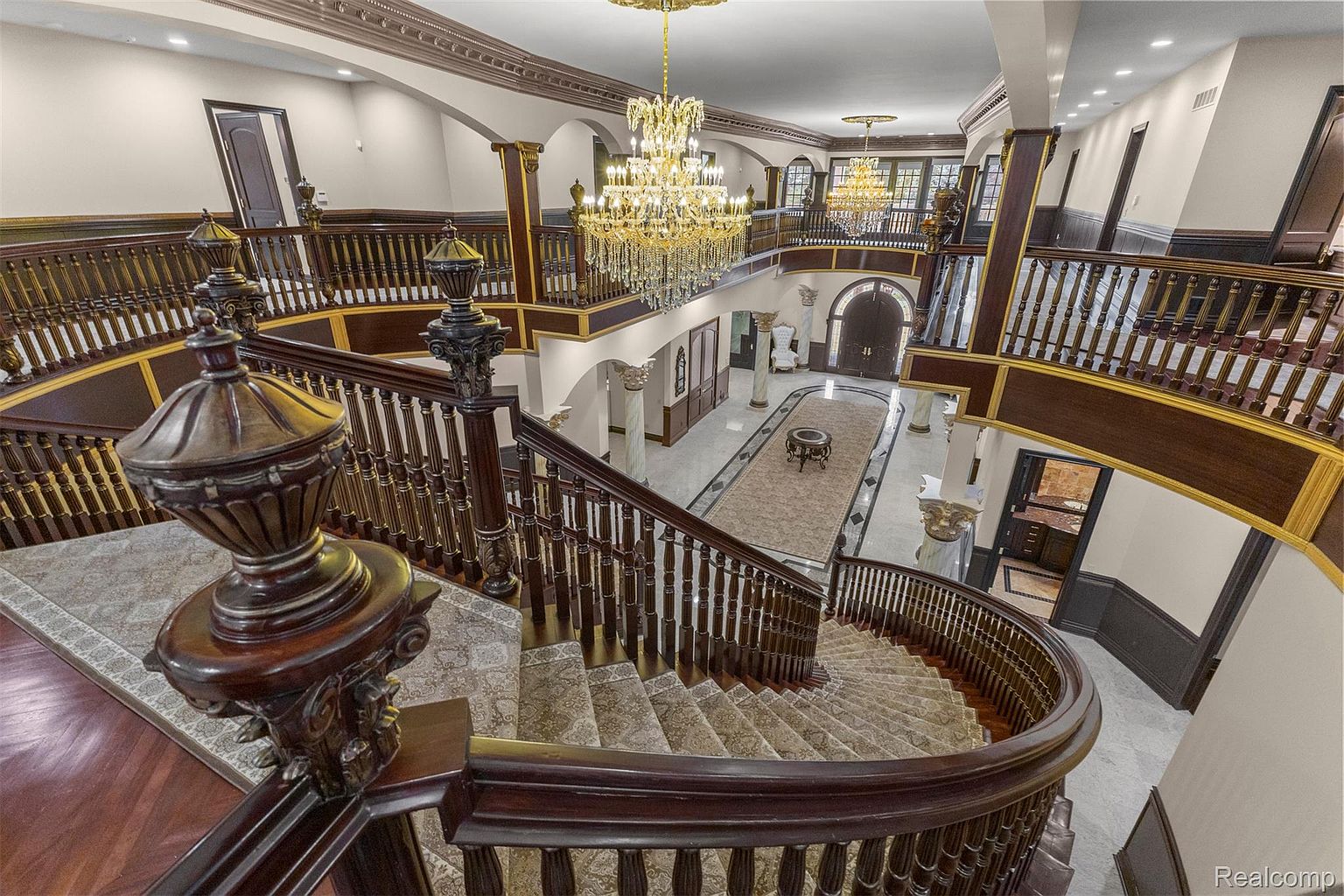 This grand interior shot showcases a luxurious foyer with a sweeping staircase, ornate dark wood railings, and a large crystal chandelier. The space features marble flooring, decorative columns, and a second-floor balcony overlooking the entrance, creating an opulent and impressive first impression. The perspective is from the top of the stairs looking down.