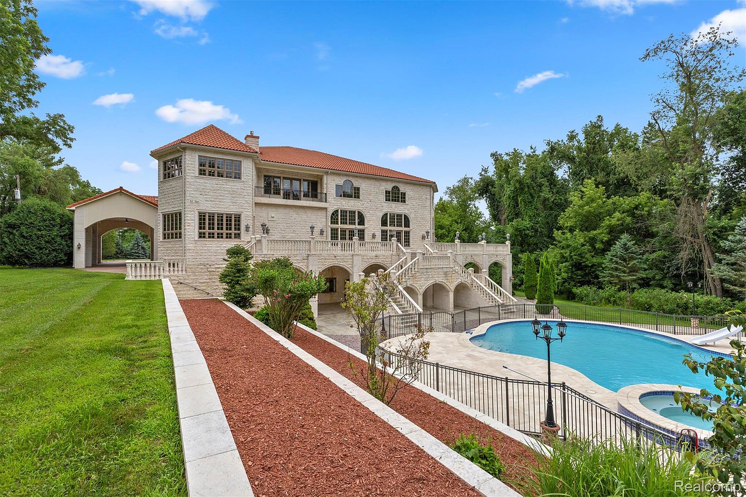 This image showcases the rear exterior of a grand, multi-story residence with a Mediterranean-inspired design. Notable features include a terracotta tile roof, light-colored stone facade, arched walkways, and multiple balconies. A luxurious swimming pool with a spa is visible in the foreground, surrounded by manicured landscaping and wrought-iron fencing, enhancing the property's appeal as a high-end estate.