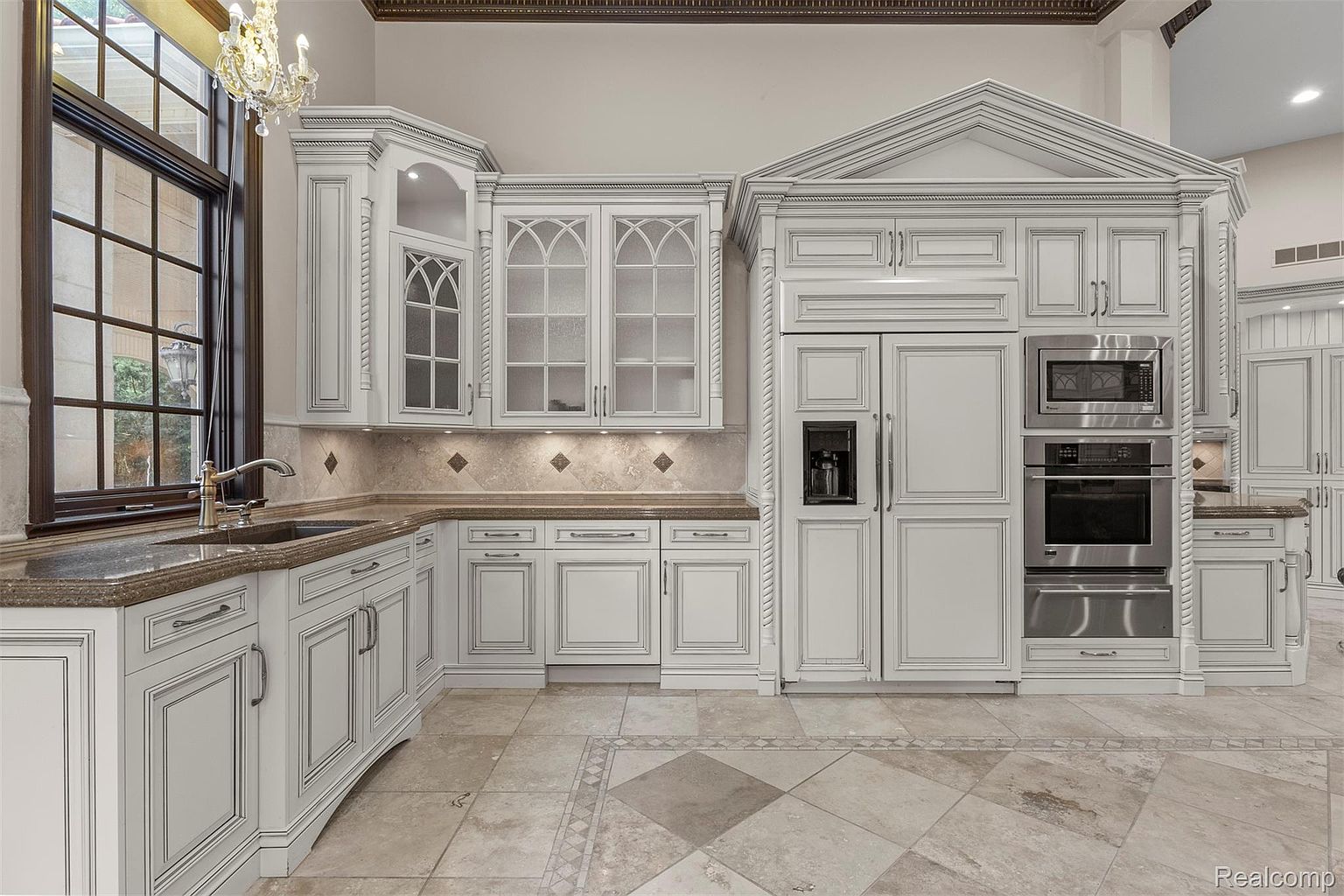 This is a well-lit kitchen featuring custom white cabinetry with detailed trim, granite countertops, and stainless steel appliances. The kitchen has a traditional design with a large window providing natural light and a decorative tile floor. The overall impression is one of luxury and sophistication.