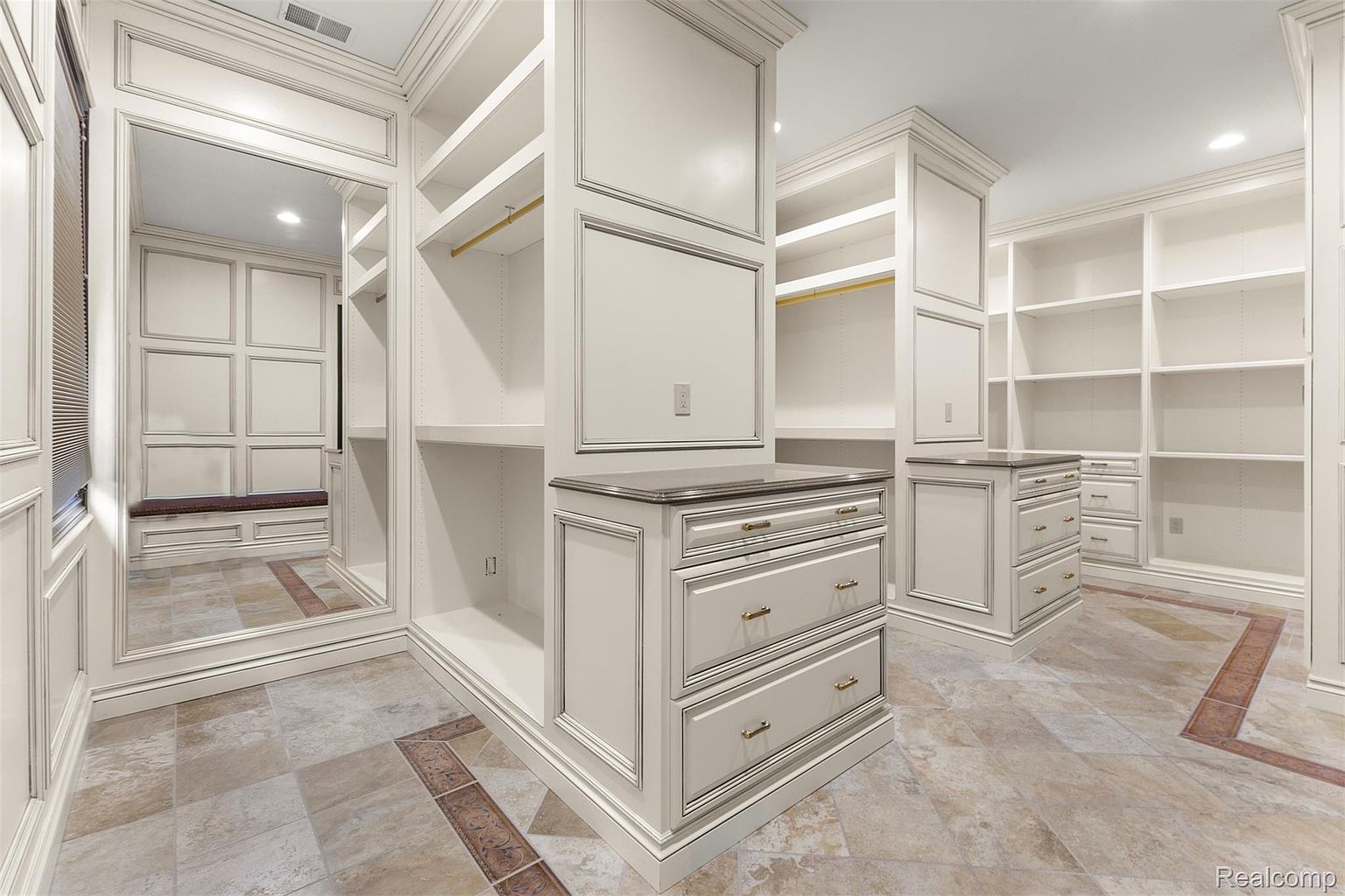 This is a spacious walk-in closet featuring custom white cabinetry with detailed trim. The closet includes shelving, drawers, and hanging rods, providing ample storage space. The flooring is a patterned tile, adding a touch of elegance to the space.