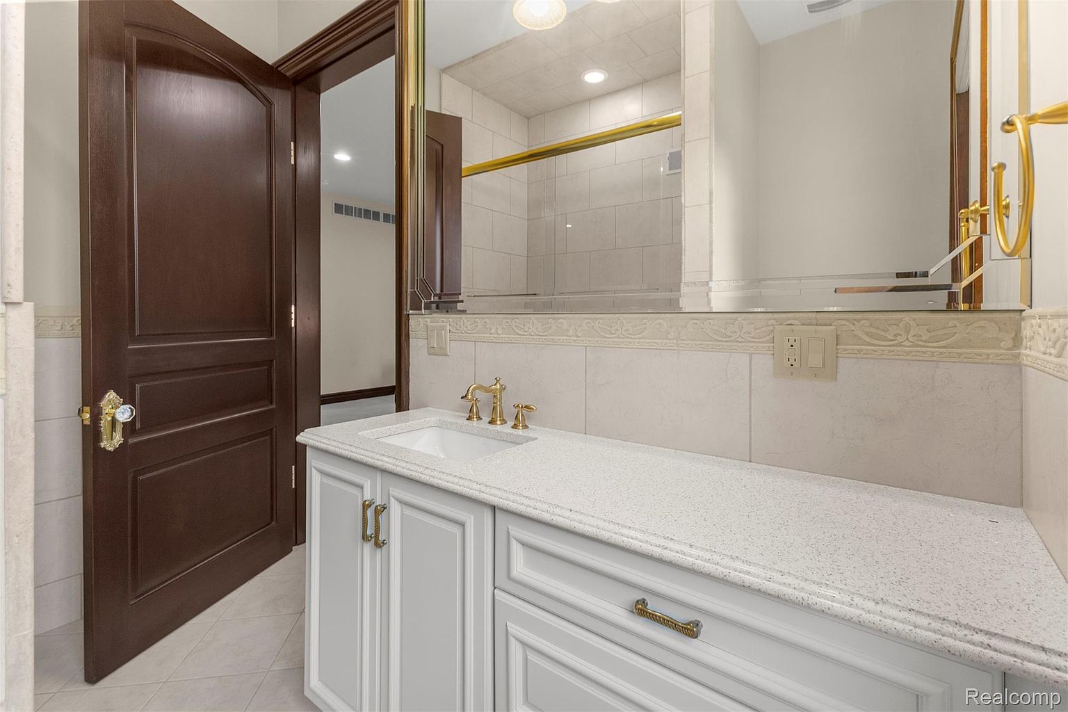 This bathroom features a white vanity with a speckled countertop and gold hardware, complemented by a matching gold faucet and fixtures. A large mirror reflects the shower area, which is tiled in a neutral tone and has a gold shower rod. A dark wood door with ornate gold hardware is partially open, adding a touch of elegance.