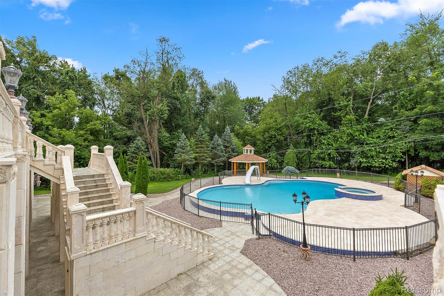 This image showcases a luxurious outdoor pool and spa area. The pool features a slide and is surrounded by a well-maintained patio and decorative fencing. The area is complemented by lush landscaping and mature trees, creating a private and serene backyard oasis. A grand staircase leads down to the pool area.