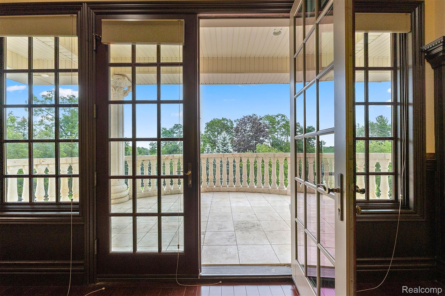 This image showcases an elegant indoor-outdoor transition with a view of a spacious balcony. The balcony features a classic balustrade and overlooks lush greenery under a clear sky. The interior space is framed by dark wood-trimmed windows and doors, adding a touch of sophistication to the scene.