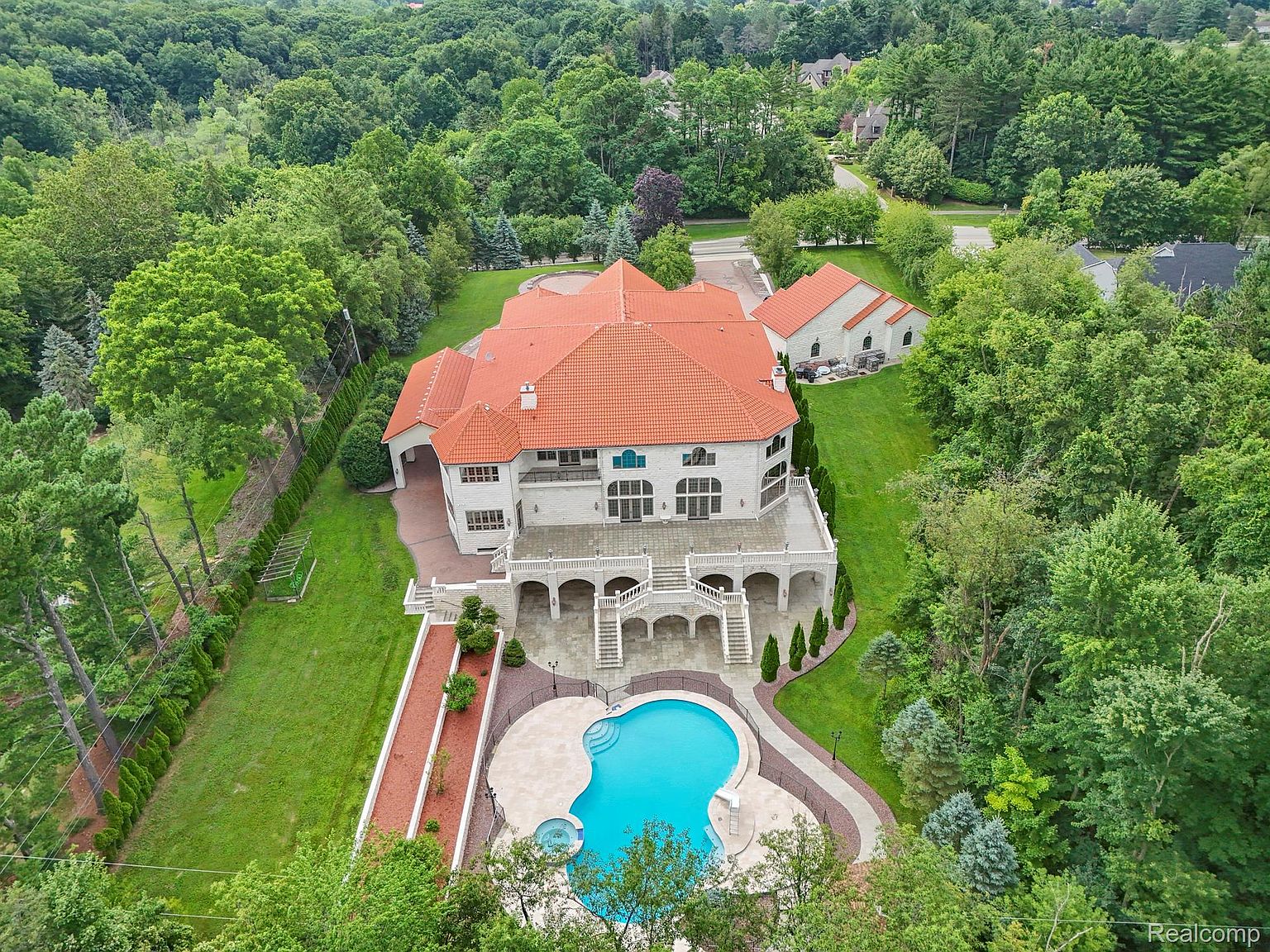 This aerial view showcases a grand estate nestled amidst lush greenery. The property features a large house with a terracotta-tiled roof, a spacious patio with elegant balustrades, and a kidney-shaped swimming pool. A separate guest house or garage is also visible, adding to the property's appeal and functionality.