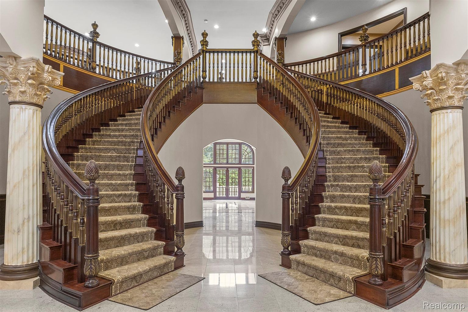 This grand foyer showcases a stunning dual staircase with ornate wooden railings and carpeted steps, creating a luxurious and inviting entrance. The staircase curves gracefully towards the upper level, flanked by elegant columns and a large window that floods the space with natural light. The polished marble flooring adds to the overall opulence and grandeur of the home.