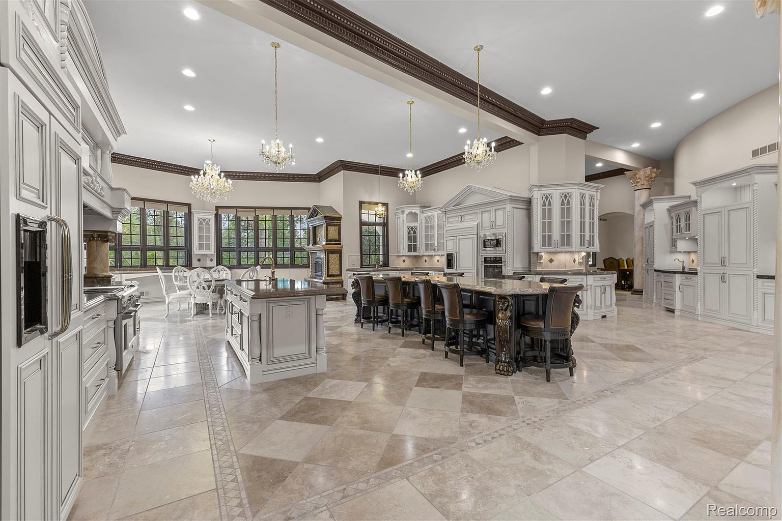 This is a spacious, luxurious kitchen featuring custom white cabinetry with detailed trim, a large central island with bar seating, and elegant chandeliers. The kitchen also includes a dining area with large windows, providing ample natural light. The overall impression is one of grandeur and sophistication, perfect for a high-end real estate listing.