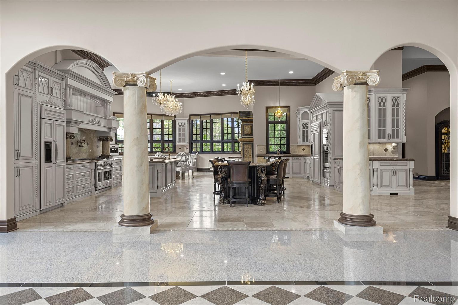 This is a grand kitchen featuring custom cabinetry, marble countertops, and a large central island with seating. The space is defined by architectural columns and arches, creating a luxurious and open feel. Chandeliers provide elegant lighting, and the overall design exudes sophistication and high-end craftsmanship.