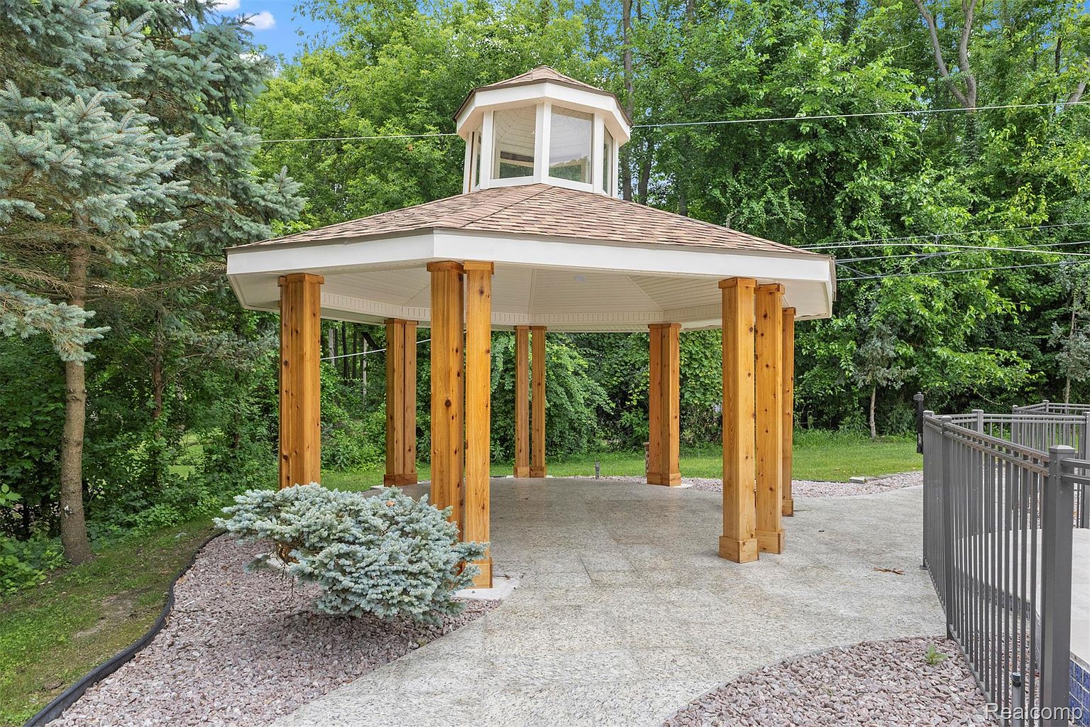 This image showcases a well-maintained yard featuring a charming gazebo with wooden pillars and a light-colored roof. The gazebo is surrounded by lush greenery and a stone pathway, creating a serene and inviting outdoor space. A decorative bush adds a touch of elegance to the landscape.