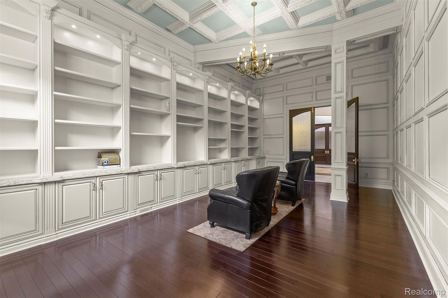 This is an interior shot of a luxurious home office or study. The room features custom built-in bookshelves with white cabinetry and marble countertops, dark hardwood floors, and paneled walls. A chandelier hangs from the coffered ceiling, and two leather armchairs sit on a rug in the center of the room, creating a sophisticated and inviting space.