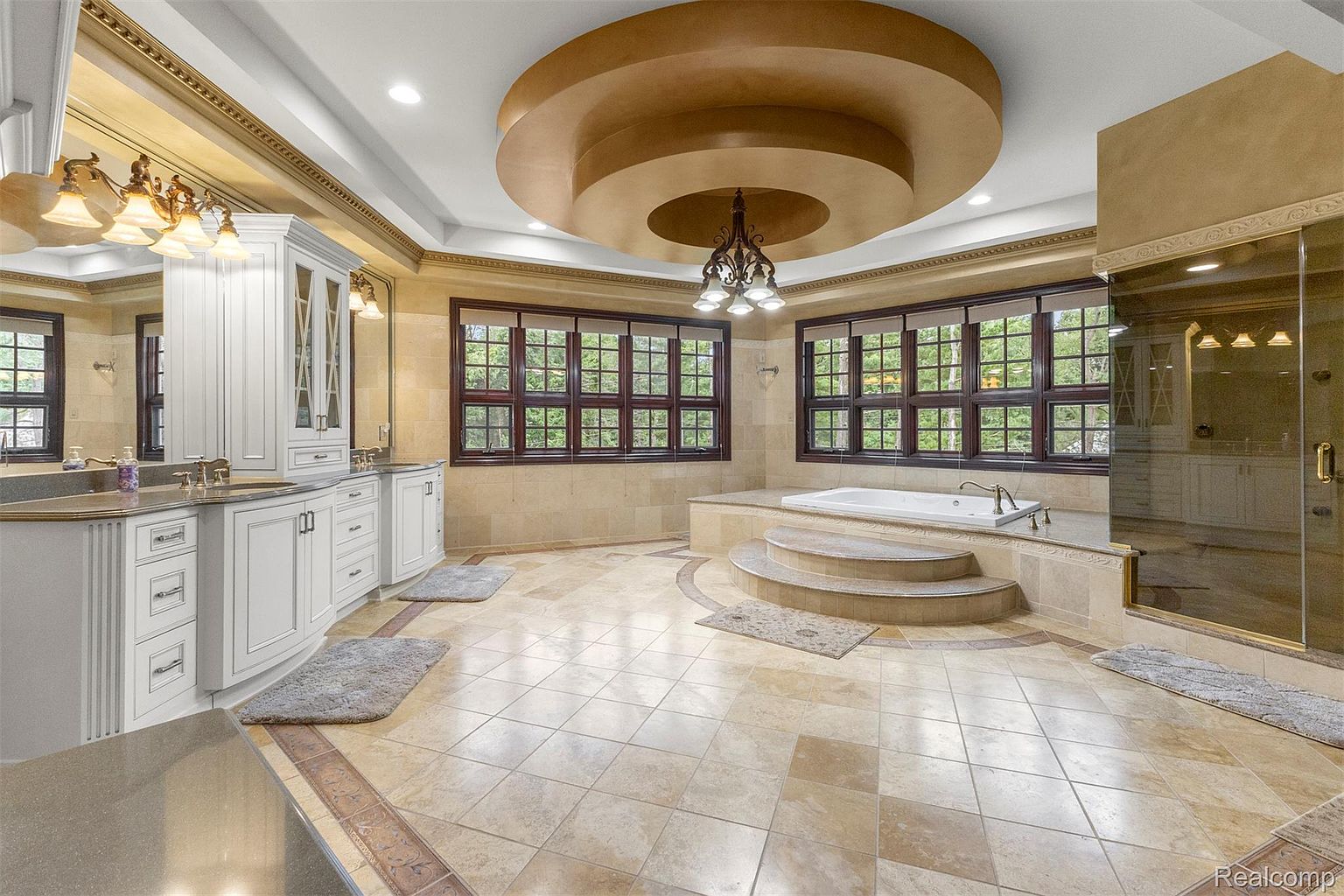 This is a luxurious primary bathroom featuring a large jacuzzi tub surrounded by steps, a glass-enclosed shower, and a double vanity with white cabinetry. The room is illuminated by a chandelier and recessed lighting, and the flooring is a light-colored tile with decorative accents. The overall impression is one of opulence and relaxation.