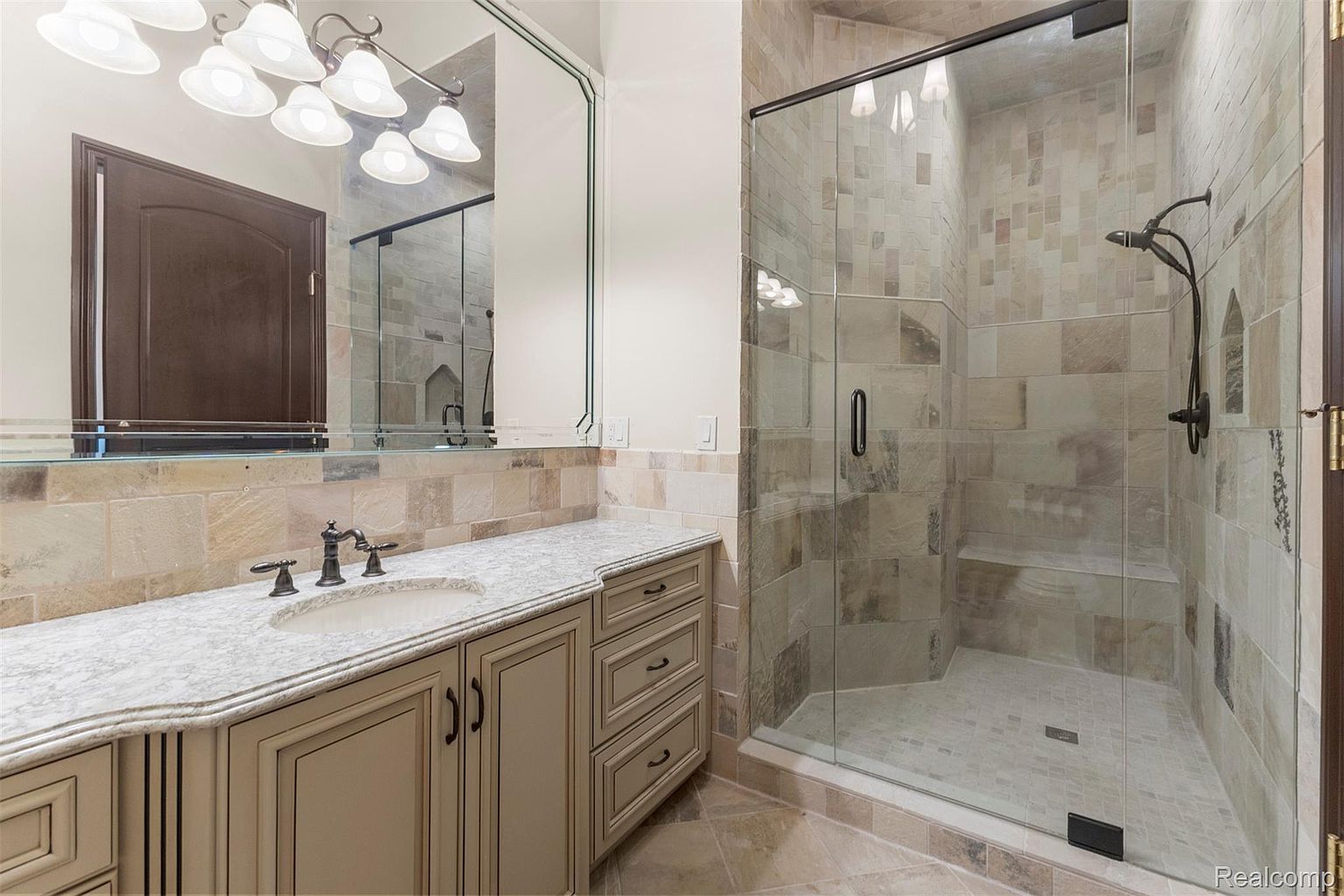 This is a well-appointed primary bathroom featuring a vanity with a marble countertop, a sink, and dark bronze fixtures. A large mirror hangs above the vanity, reflecting the room's lighting and a glimpse of the shower. The shower is enclosed in glass and features tiled walls and a built-in bench, creating a luxurious and spa-like atmosphere.