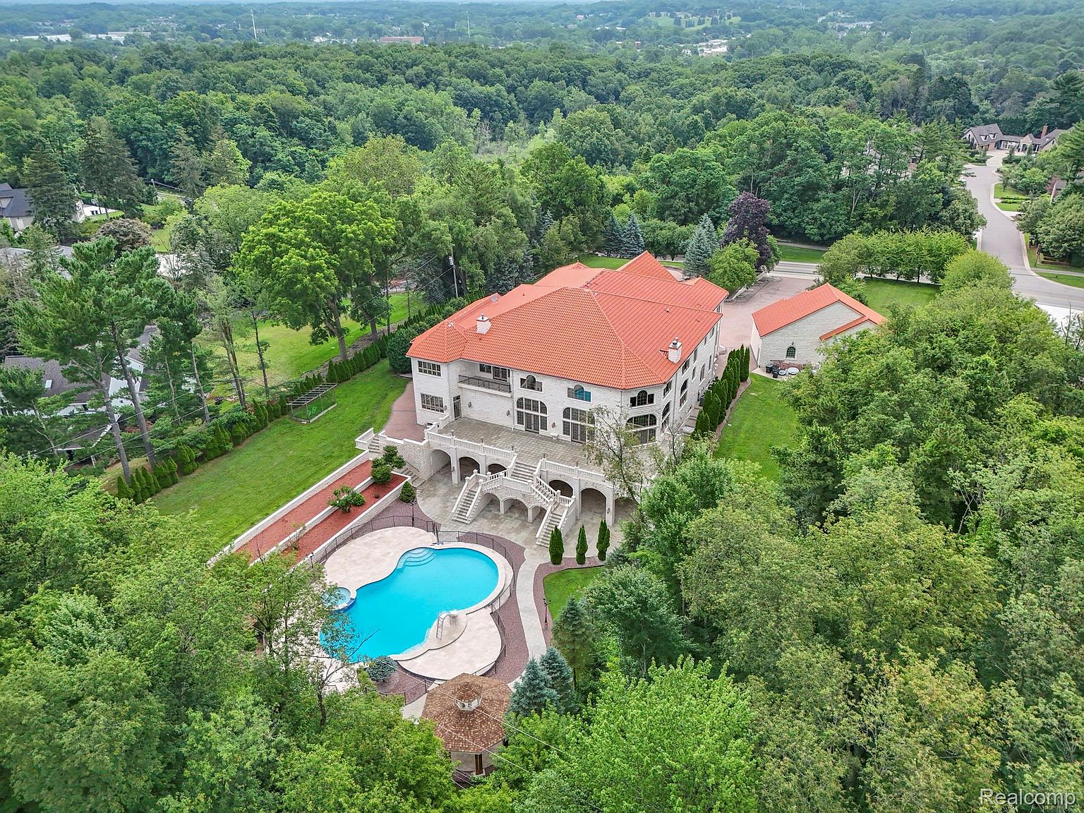 This aerial view showcases a grand estate nestled amidst lush greenery. The property features a large house with a red tile roof, a swimming pool with a unique shape, and meticulously landscaped grounds. The overall impression is one of luxury, privacy, and expansive outdoor living.
