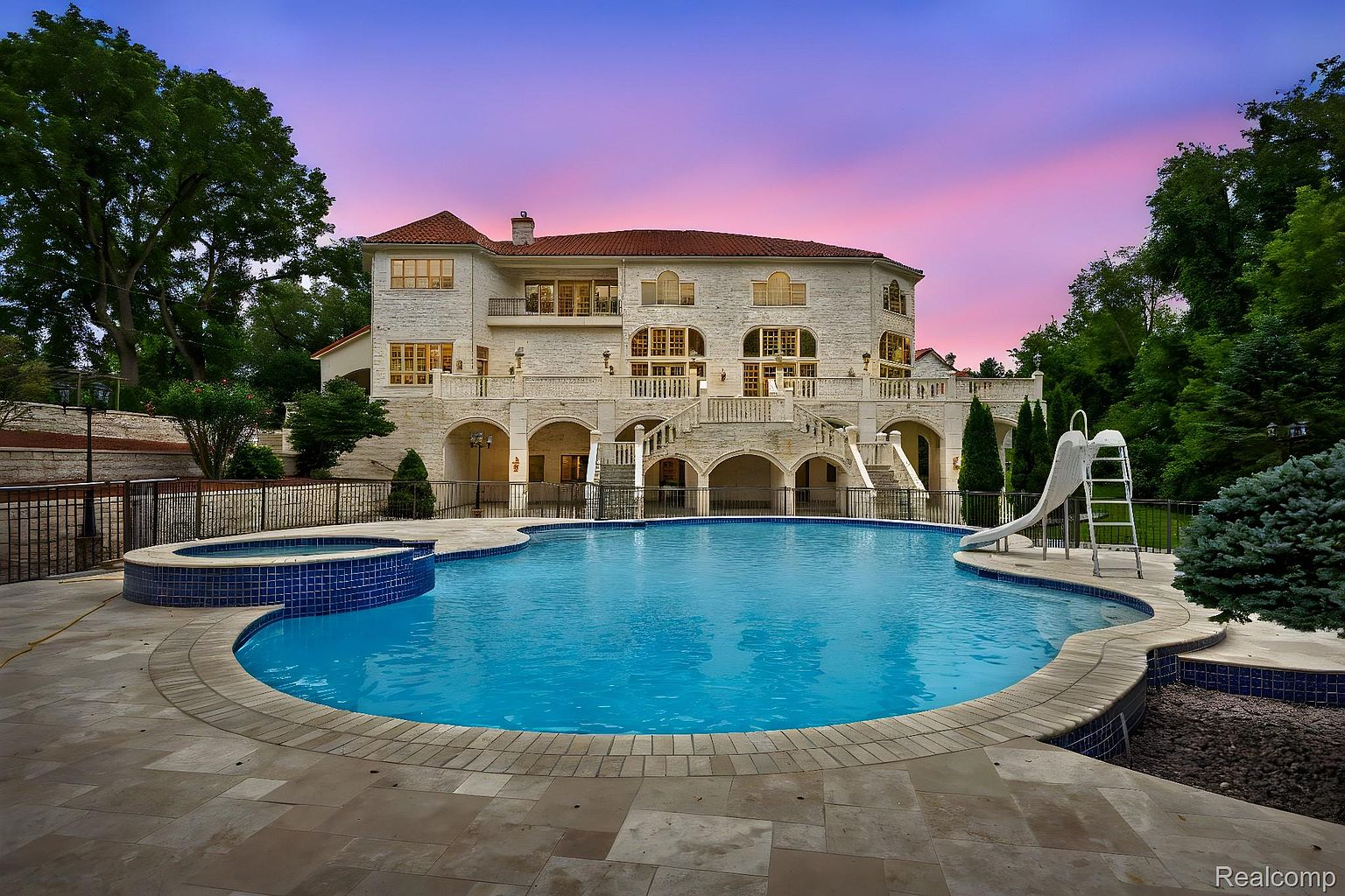 This image showcases a luxurious backyard featuring a large, kidney-shaped swimming pool with a built-in spa. The pool is surrounded by a stone patio and overlooks a grand, multi-story house with arched walkways and balconies. A slide adds a playful touch to the pool area, enhancing the property's appeal for families.