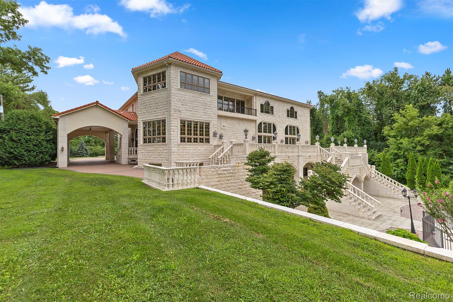 This is a front exterior view of a grand, multi-level stone house with a red tile roof. The house features multiple windows, balconies, and elaborate staircases leading to the main entrance. A covered carport is attached to the side of the house, and a well-manicured lawn slopes gently towards the viewer, creating an inviting and luxurious curb appeal.