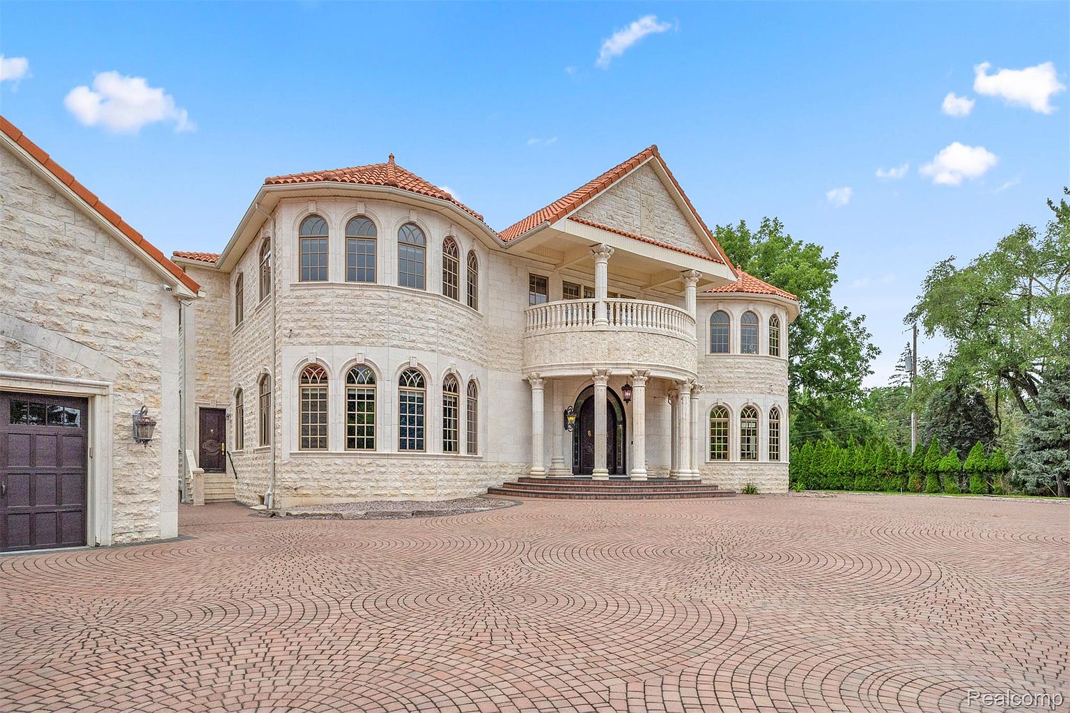 This is a front exterior view of a grand, luxurious home. The house features a light-colored stone facade, a red tile roof, and multiple arched windows. A circular driveway made of red brick pavers leads up to the entrance, which is adorned with columns and a balcony. The overall impression is one of elegance and opulence.
