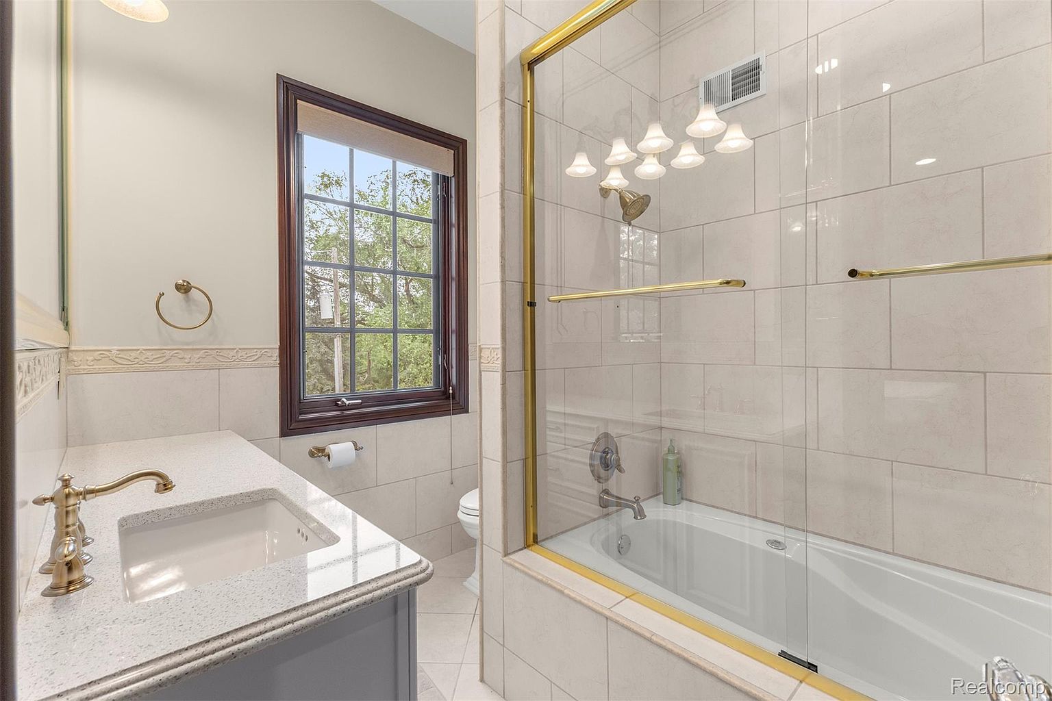 This bathroom features a white bathtub with a glass enclosure and gold trim, complemented by light beige tiled walls. A vanity with a white countertop and brass faucet is visible, along with a window providing natural light. The overall style is traditional and well-maintained.