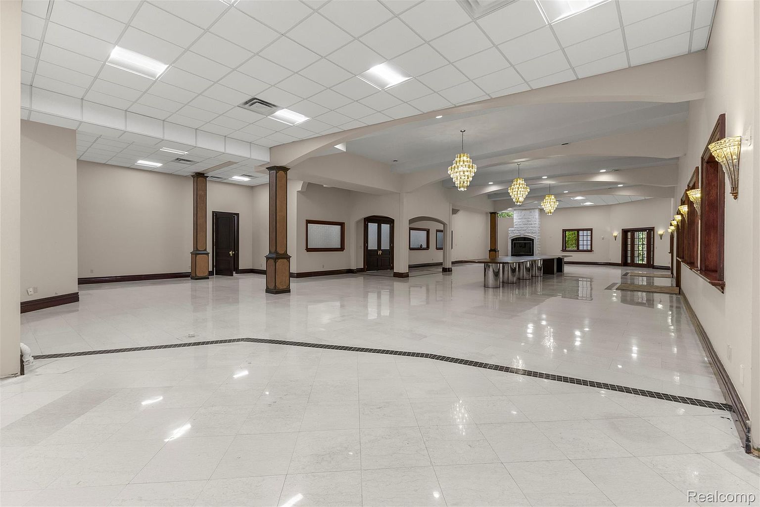 This is a spacious living room featuring a white tile floor with dark accents, multiple pillars with wood trim, and recessed lighting in the ceiling. The room has a neutral color palette with dark wood accents on the doors and windows, and several chandeliers hang from the ceiling. The room appears to be designed for large gatherings or events.