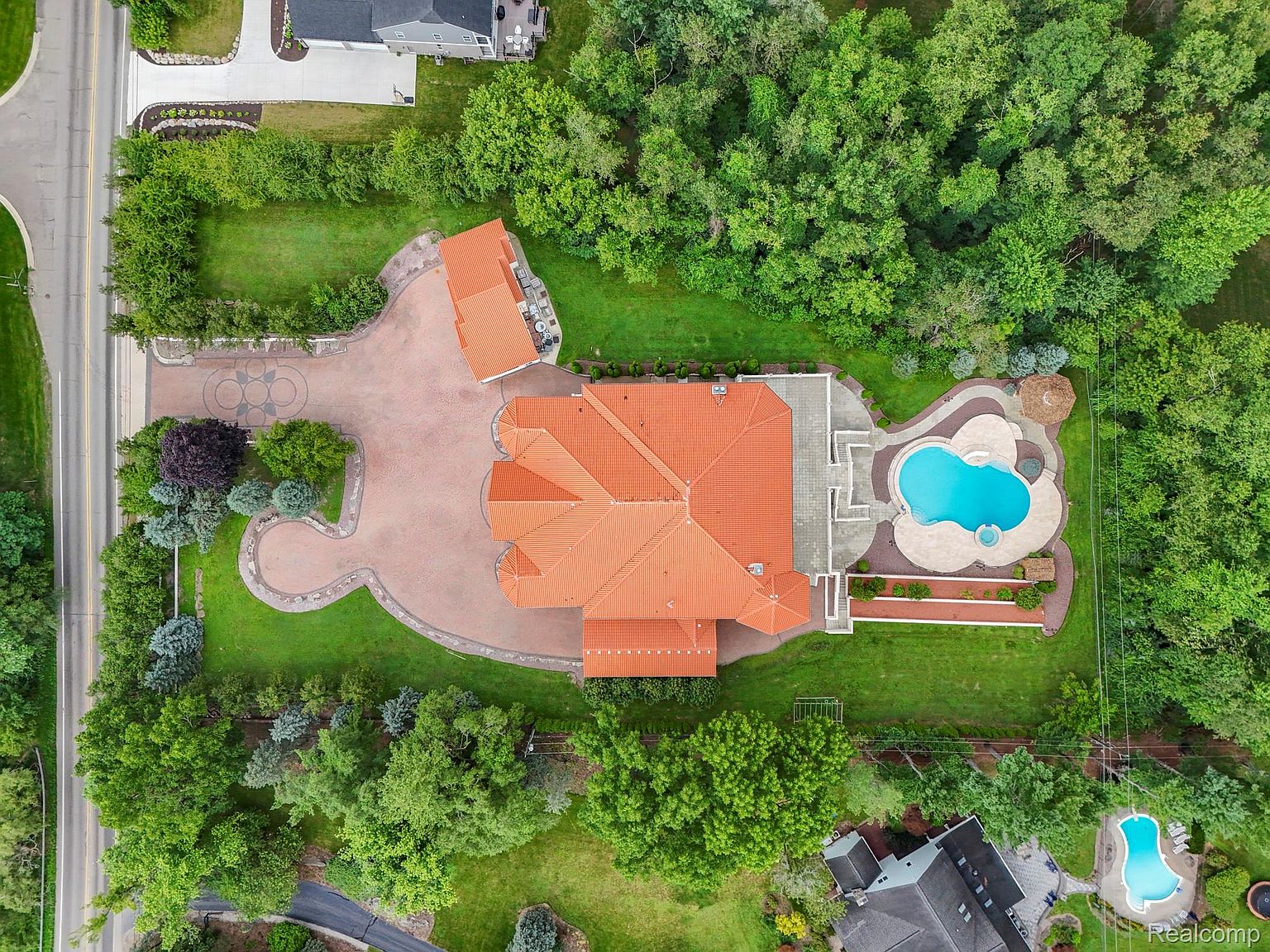This aerial shot showcases a luxurious estate with a sprawling red-tiled roof, a meticulously landscaped yard, and a private pool area. The property features a circular driveway leading to the main house, surrounded by lush greenery and mature trees, creating a sense of privacy and exclusivity. The pool area includes a unique clover-shaped pool and a spa, perfect for relaxation and entertainment.