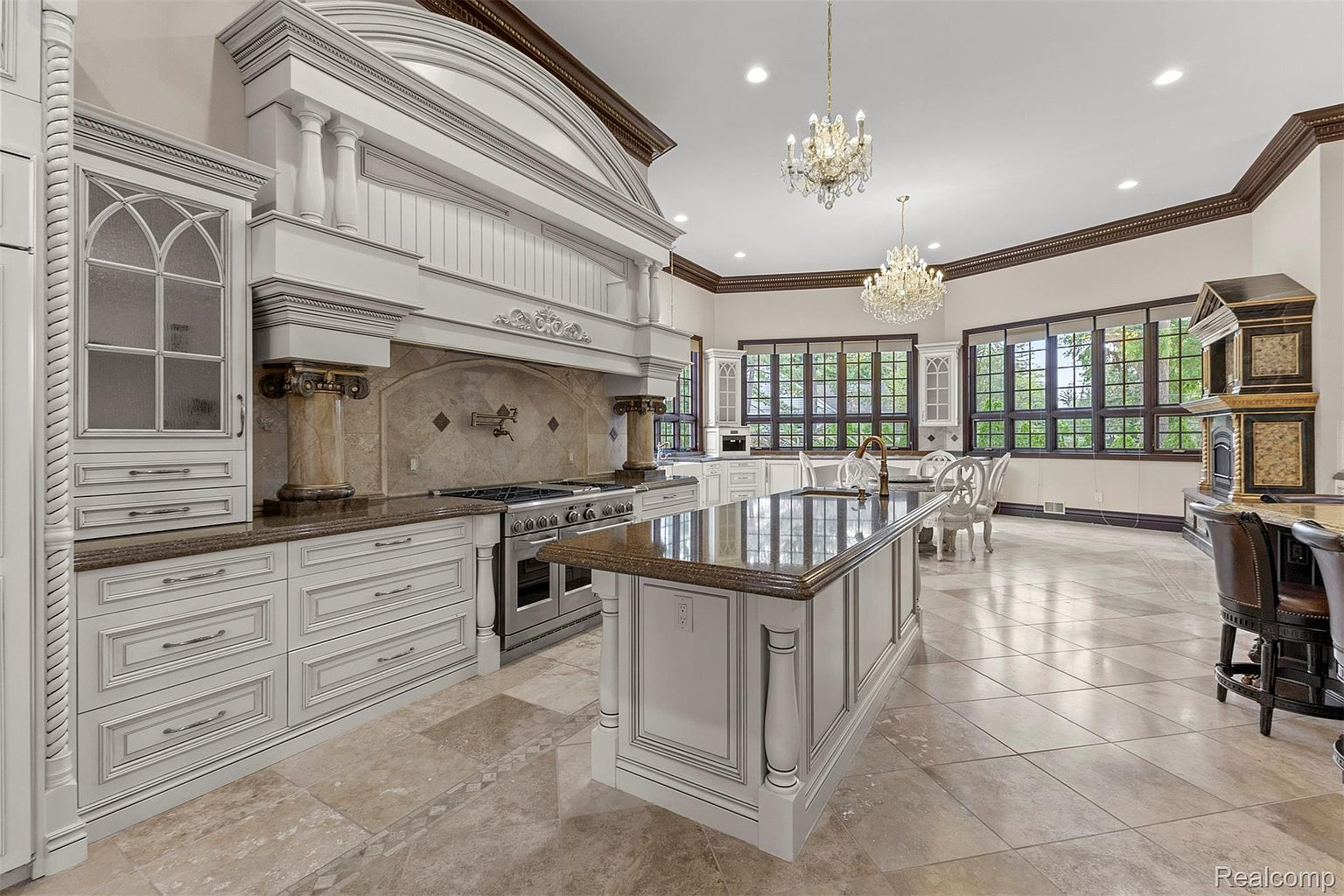 This is a luxurious kitchen featuring custom white cabinetry with detailed trim, a large central island with a dark countertop, and high-end stainless steel appliances. The kitchen is illuminated by two ornate chandeliers, and the flooring is a patterned tile. The overall impression is one of elegance and sophistication.