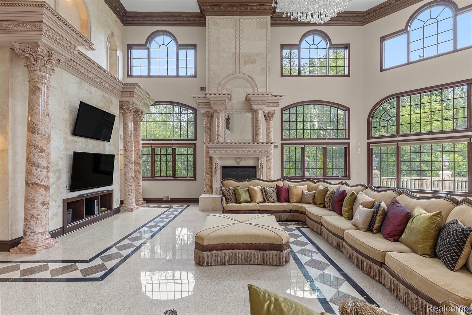 This grand living room features soaring ceilings, marble columns, and a large fireplace, creating a luxurious and opulent atmosphere. The expansive windows offer ample natural light, highlighting the detailed architectural features and the elegant, oversized sectional sofa. The polished marble floors and the intricate ceiling details add to the room's sophisticated charm.