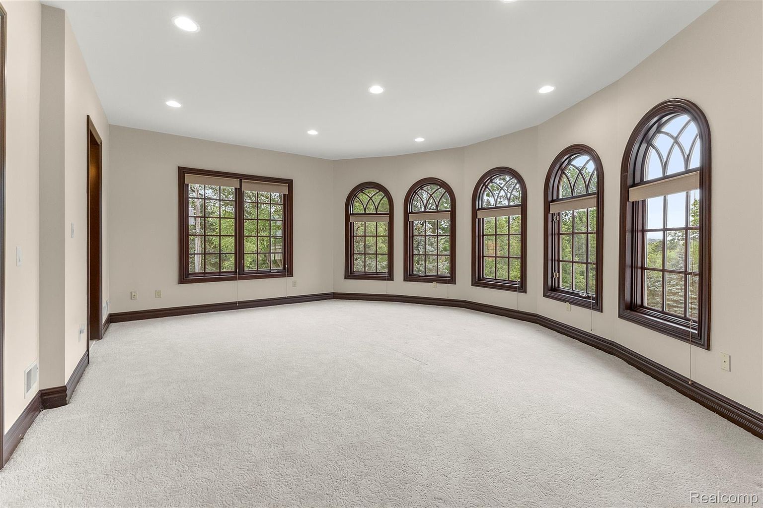 This is a spacious room with white carpet and neutral-colored walls, featuring a series of arched windows with dark wood trim that provide ample natural light. The room has a clean and minimalist aesthetic, with dark wood trim along the base of the walls, creating a sense of elegance and sophistication. Recessed lighting is present throughout the ceiling.