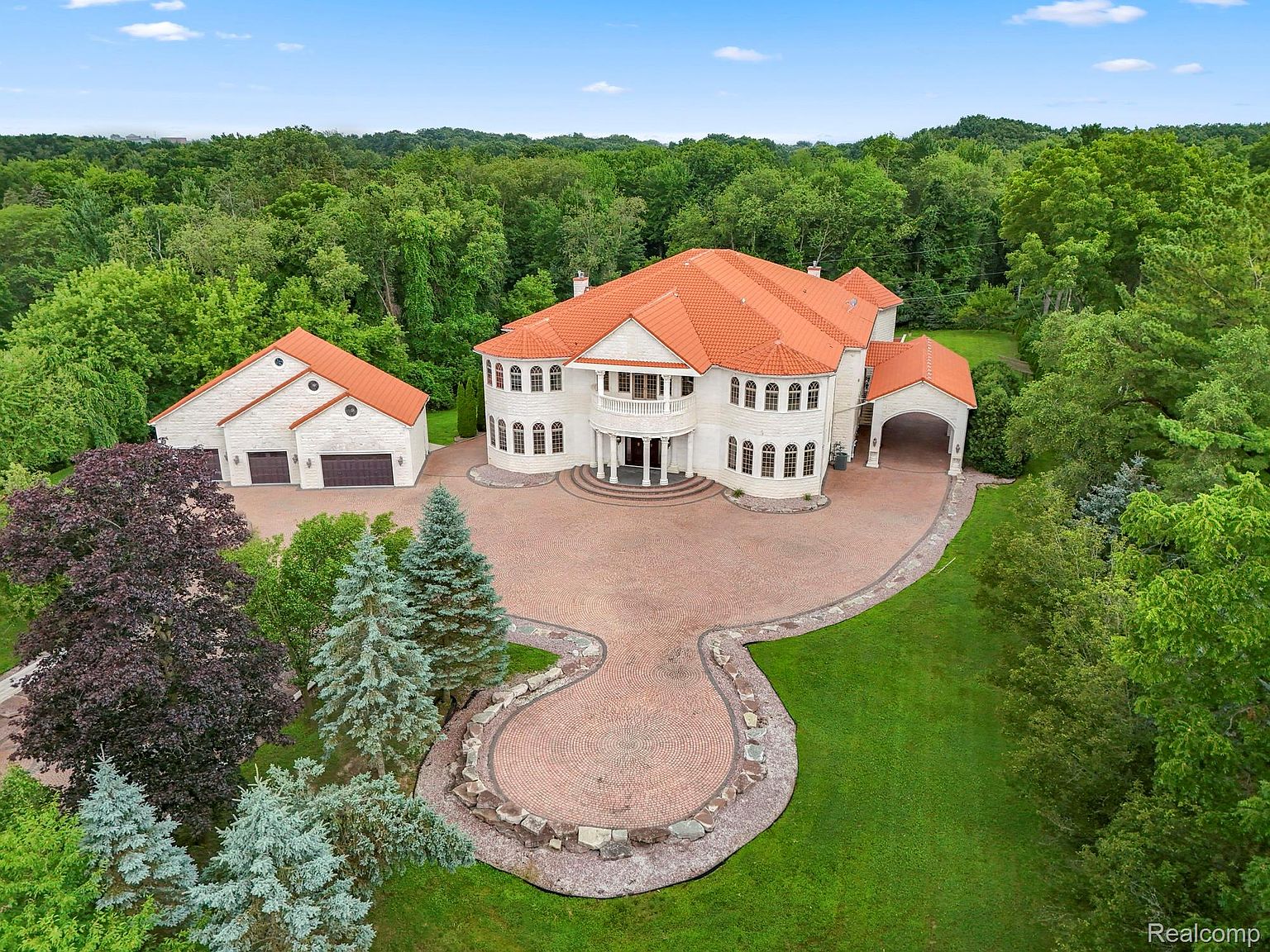 This aerial view showcases a grand estate with a sprawling red-tiled roof, a circular driveway, and meticulously landscaped grounds. The main house features a light-colored facade with arched windows and a prominent balcony, complemented by a detached garage with matching architectural details. Lush greenery surrounds the property, providing privacy and a sense of seclusion.