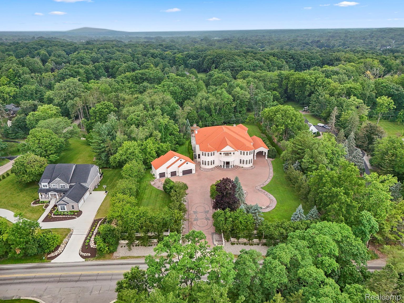 This aerial view showcases a grand estate with a terracotta-tiled roof, set amidst lush greenery. A long, patterned driveway leads to the main house and a detached garage, both featuring matching roof tiles. The surrounding landscape is densely wooded, providing privacy and a sense of seclusion, while a neighboring house is visible to the left.