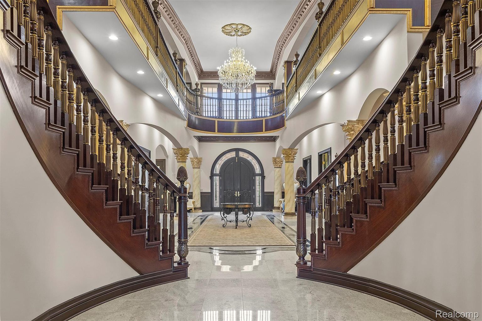 This grand foyer features dual curved staircases with dark wood and gold accents, leading to a second-floor balcony overlooking the entrance. A large crystal chandelier hangs from the ceiling, illuminating the marble flooring and a central table on a decorative rug. The space is framed by arched doorways and ornate columns, creating a luxurious and impressive first impression.