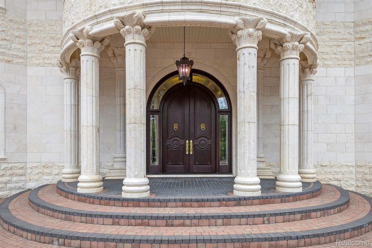 This image showcases the grand entryway of a luxurious home, featuring a set of ornate double doors framed by classical columns and an arched portico. The entrance is elevated by a series of curved steps, creating a welcoming and impressive approach. The architectural details and high-end materials suggest a property of significant value and refined taste.
