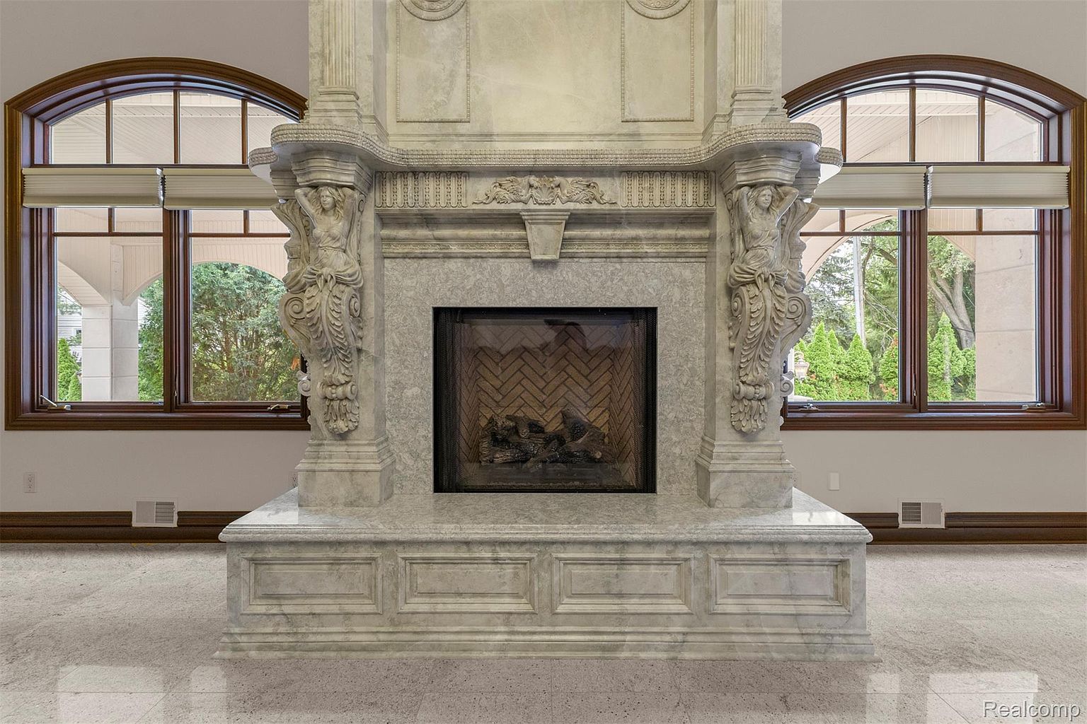 The image showcases a grand, ornate fireplace constructed from marble or a similar stone material. The fireplace features intricate carvings, including sculpted figures on either side, and is flanked by arched windows that provide natural light. The overall impression is one of luxury and classical elegance, suggesting a high-end property.