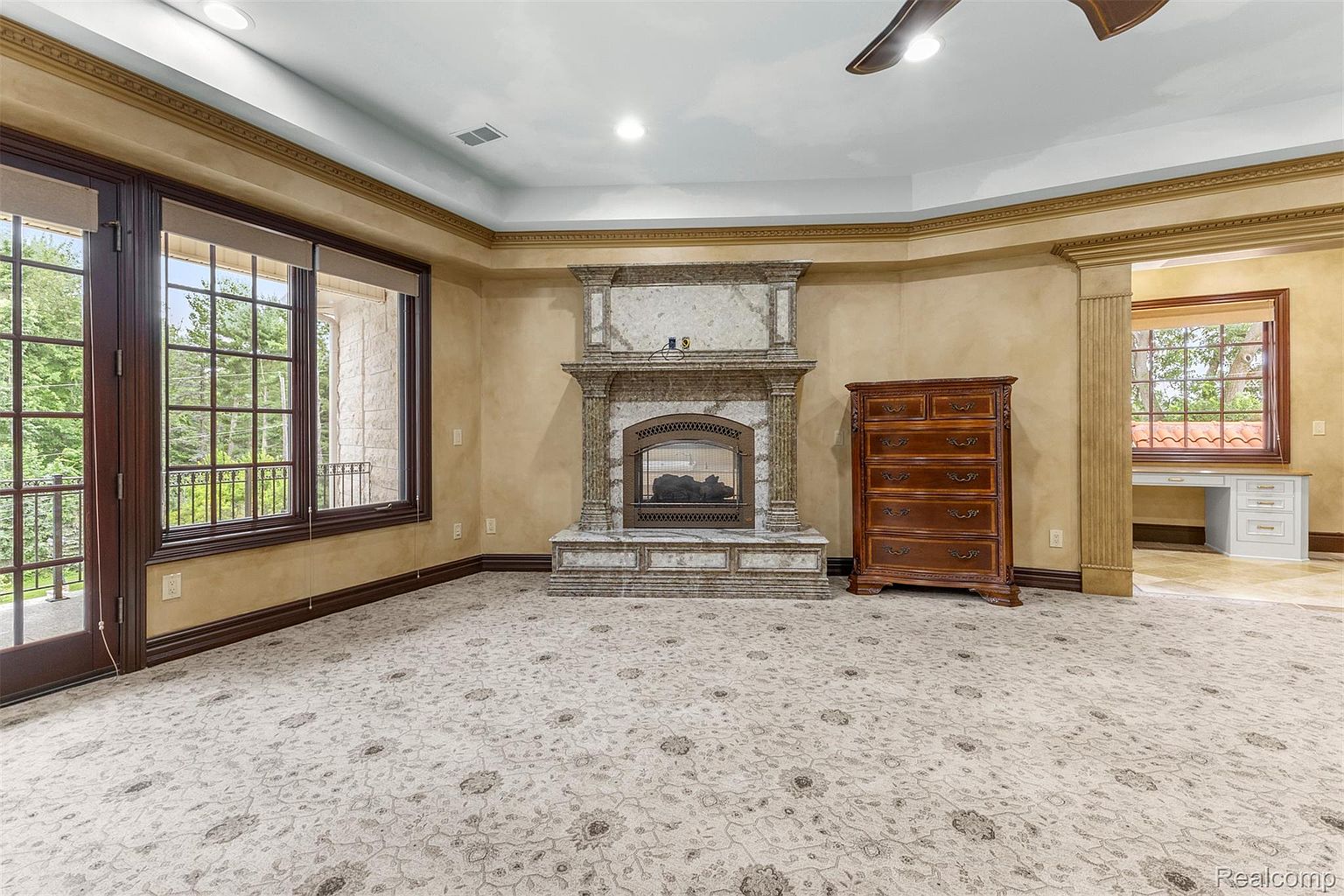 This is a spacious primary bedroom featuring a decorative fireplace with a marble surround, a large window with a view of the outdoors, and a wooden dresser. The room has a neutral color palette with beige walls and a patterned carpet. The overall impression is luxurious and comfortable, suggesting a relaxing retreat.