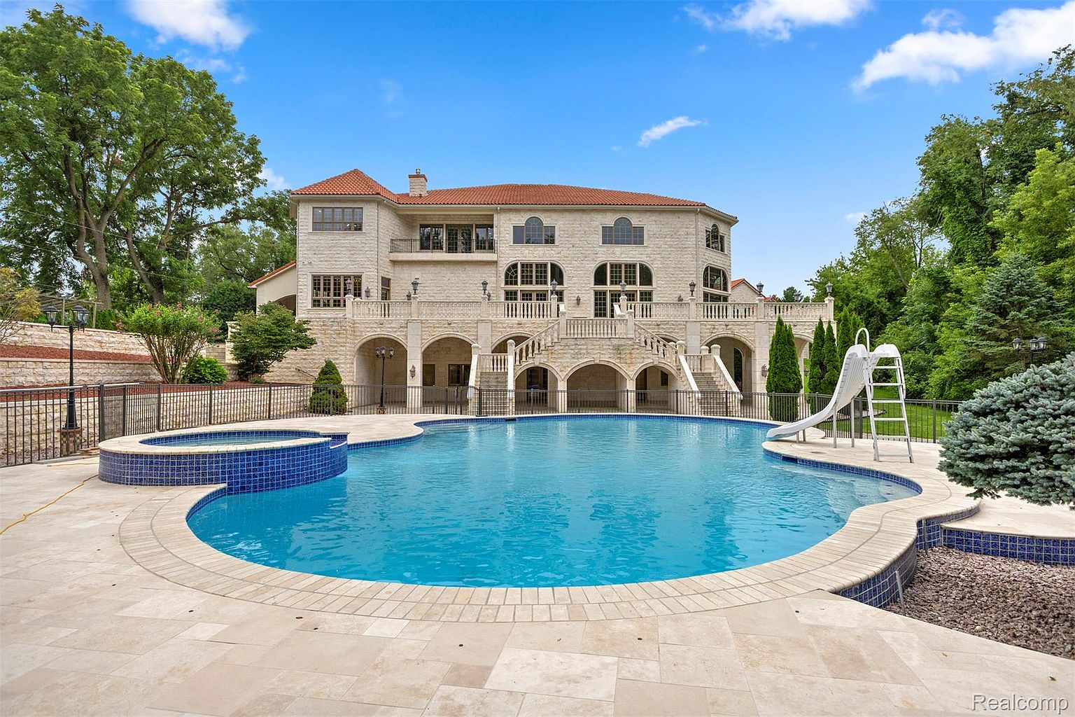 This image showcases a luxurious backyard featuring a large, kidney-shaped swimming pool with a built-in spa. The pool is surrounded by a stone patio and overlooks a grand, multi-story house with arched architectural details and a red tile roof. A water slide adds a playful element to the scene, creating an inviting and upscale outdoor living space.