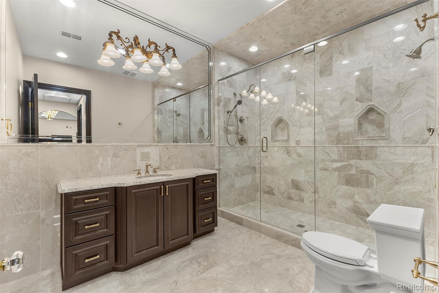 This is a primary bathroom featuring a dark wood vanity with a light countertop, a large mirror, and a glass-enclosed shower. The walls and floor are covered in light-colored tile, creating a bright and clean space. A toilet is visible to the right of the shower, and the overall style is luxurious and well-maintained.
