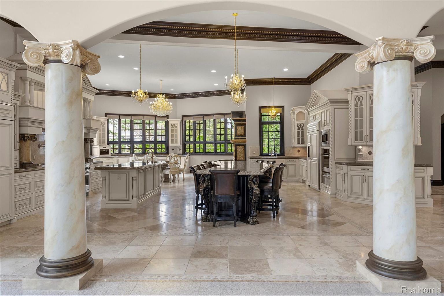 This is a grand kitchen featuring a large central island with seating, surrounded by custom cabinetry and countertops. The room is illuminated by chandeliers and natural light from the large windows. Marble columns and detailed ceiling trim add to the luxurious feel of the space.