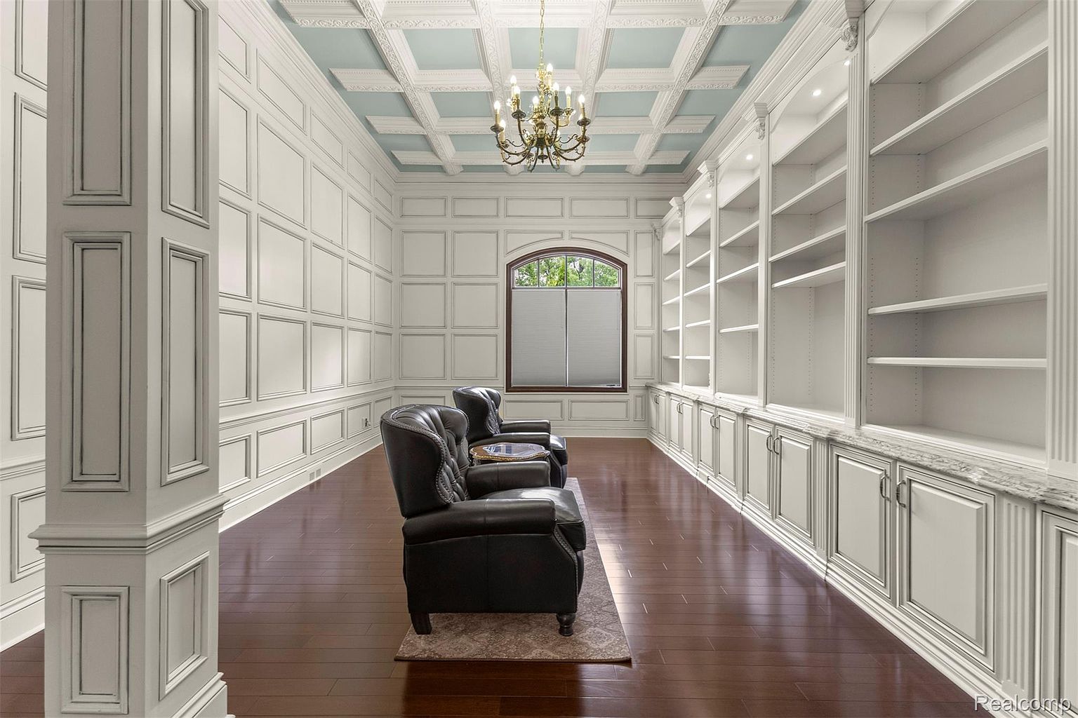 This is an interior shot of a luxurious home office or study. The room features dark hardwood floors, white paneled walls, a coffered ceiling with a chandelier, and built-in bookshelves. Two leather armchairs face a window, creating a sophisticated and comfortable space for work or relaxation.