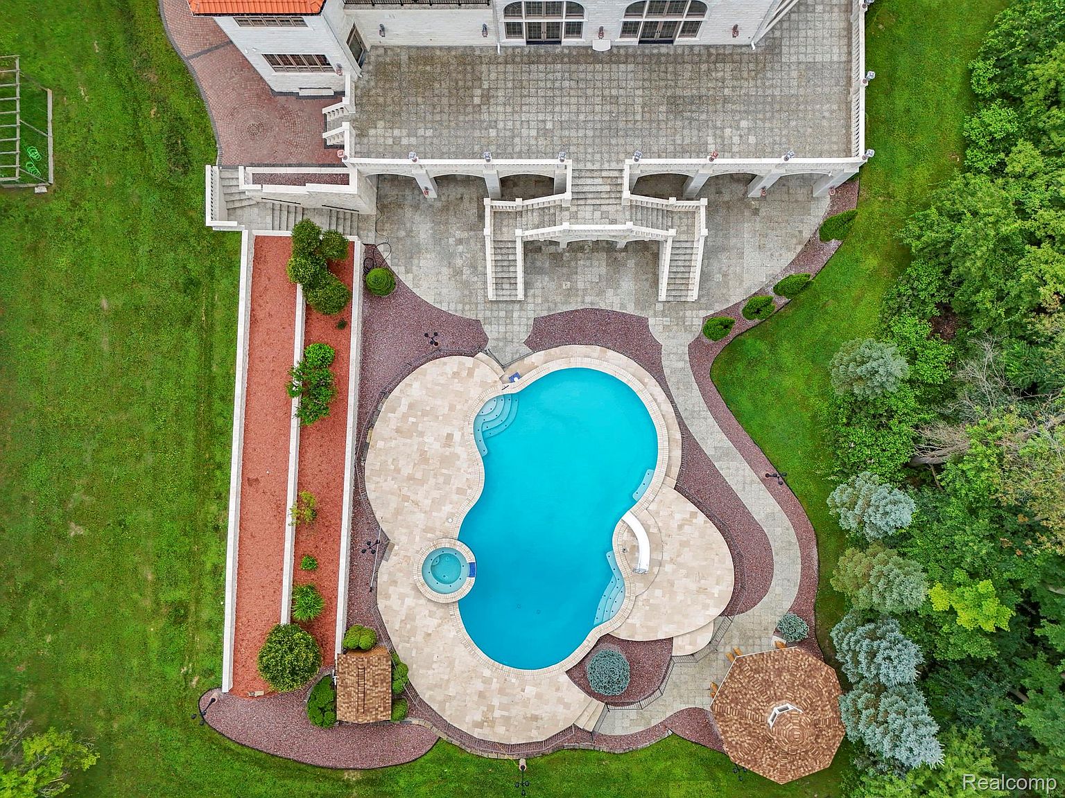 This aerial shot showcases a luxurious backyard featuring a uniquely shaped pool with a spa, surrounded by a stone patio and meticulously landscaped gardens. The property includes a grand staircase leading from a large patio area connected to the house, with mature trees and manicured lawns adding to the estate's appeal. The overall impression is one of opulence and meticulous design.