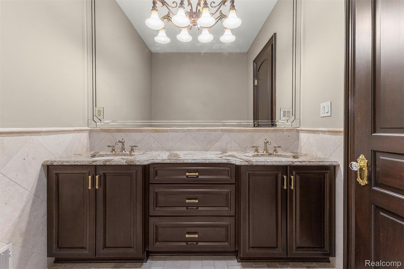 This is a well-appointed bathroom featuring a double vanity with a granite countertop and dark wood cabinetry. A large mirror reflects the vanity and a decorative chandelier, enhancing the room's elegance. The walls are adorned with light-colored tiles, contributing to a bright and clean aesthetic.