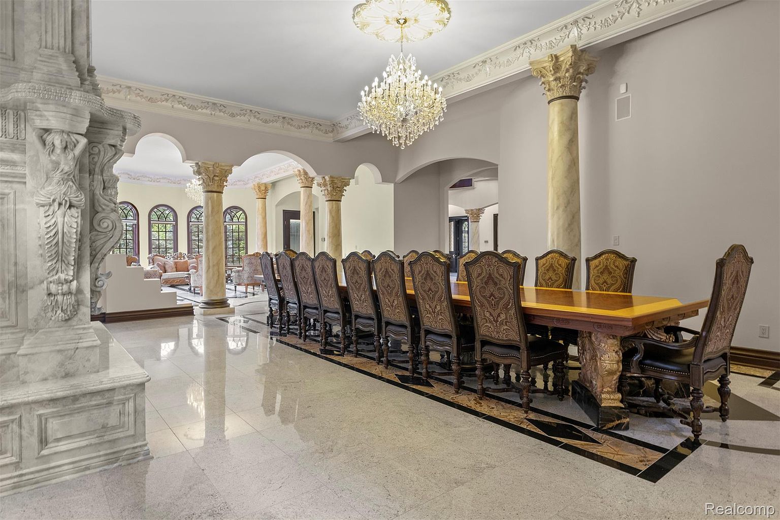 This is a grand dining room featuring a long, ornate wooden table with upholstered chairs, set on a marble floor with a decorative border. The room is adorned with marble columns, arches, and a large crystal chandelier, creating a luxurious and formal atmosphere. The perspective is from the entrance of the room, showcasing the length of the table and the architectural details.
