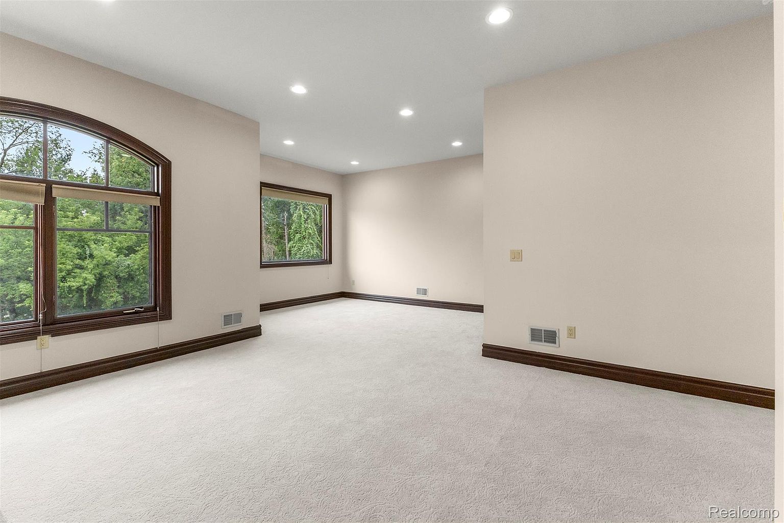 This is a spacious bedroom featuring neutral-toned walls, white carpeting, and dark wood trim. The room has two large windows that provide ample natural light and views of the surrounding greenery. Recessed lighting illuminates the room, creating a bright and inviting atmosphere.