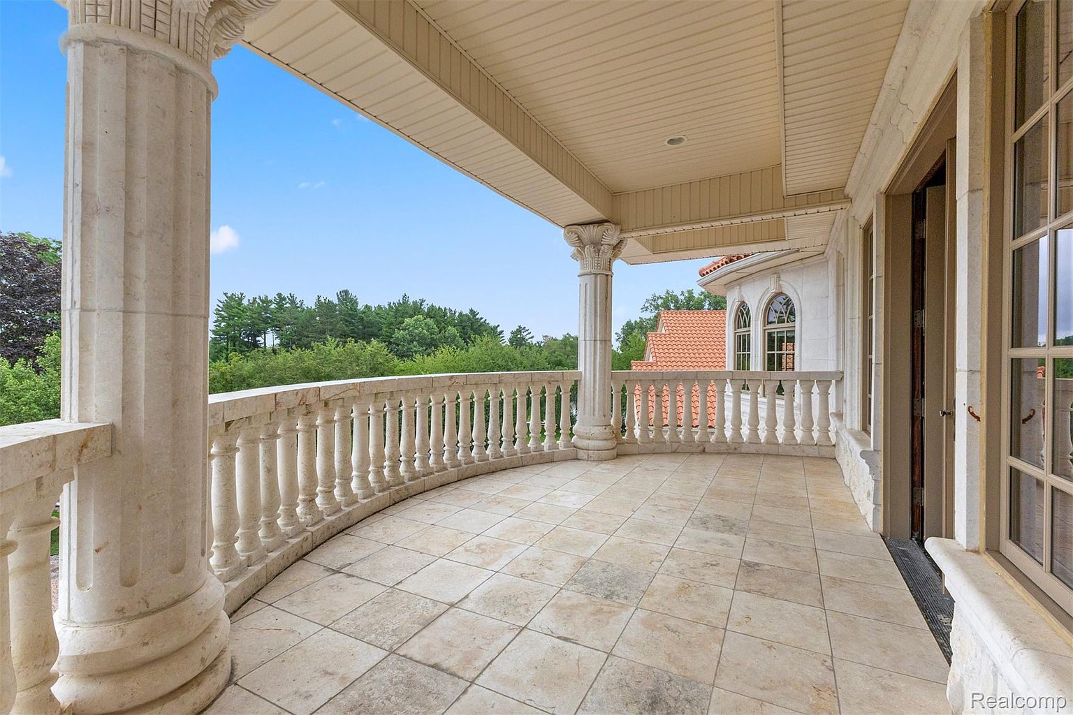This image showcases a luxurious balcony with a curved design, featuring ornate stone columns and balustrades. The flooring is composed of large, light-colored tiles, and the balcony offers a view of lush greenery and a clear blue sky. The architectural style suggests high-end craftsmanship and a focus on elegant outdoor living.