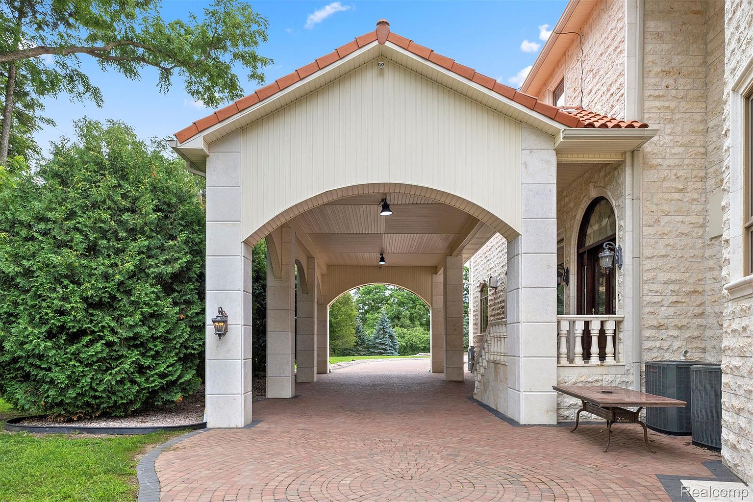 This image showcases a grand entryway featuring a covered portico with arched openings and stone columns. The driveway is paved with red brick in a circular pattern, leading to the entrance. The architectural style is luxurious and inviting, creating a strong first impression.