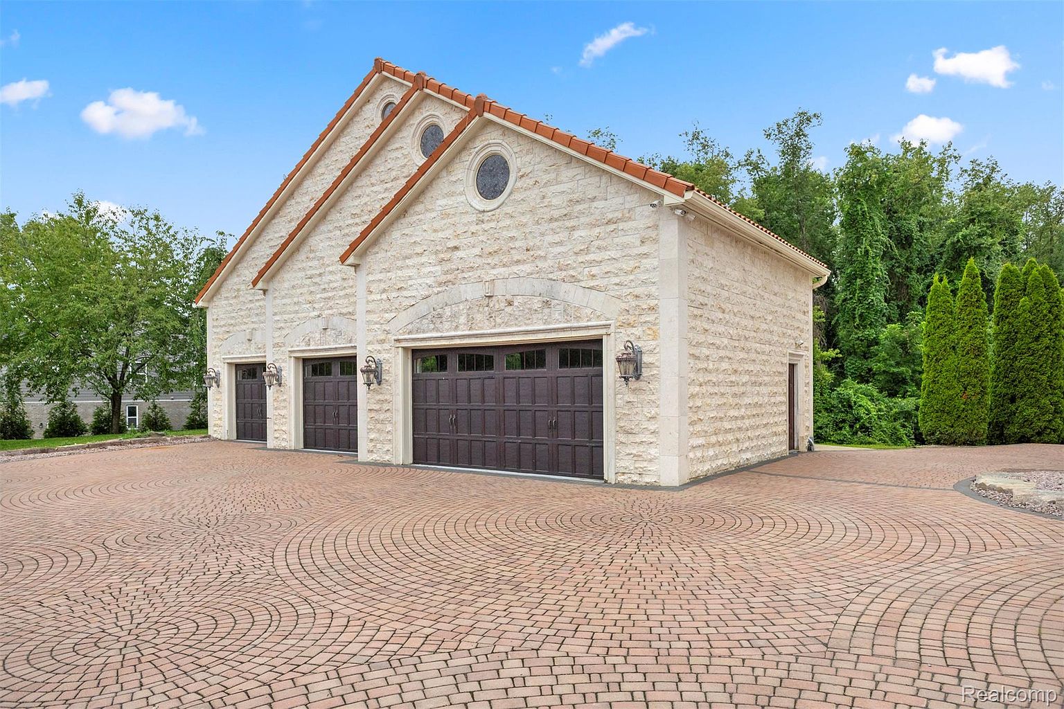 This image showcases a detached garage with three bays, constructed from light-colored stone and featuring a red tile roof. The driveway is paved with a circular brick pattern, adding to the property's curb appeal. The garage's design complements the main house, suggesting a cohesive and high-end aesthetic.