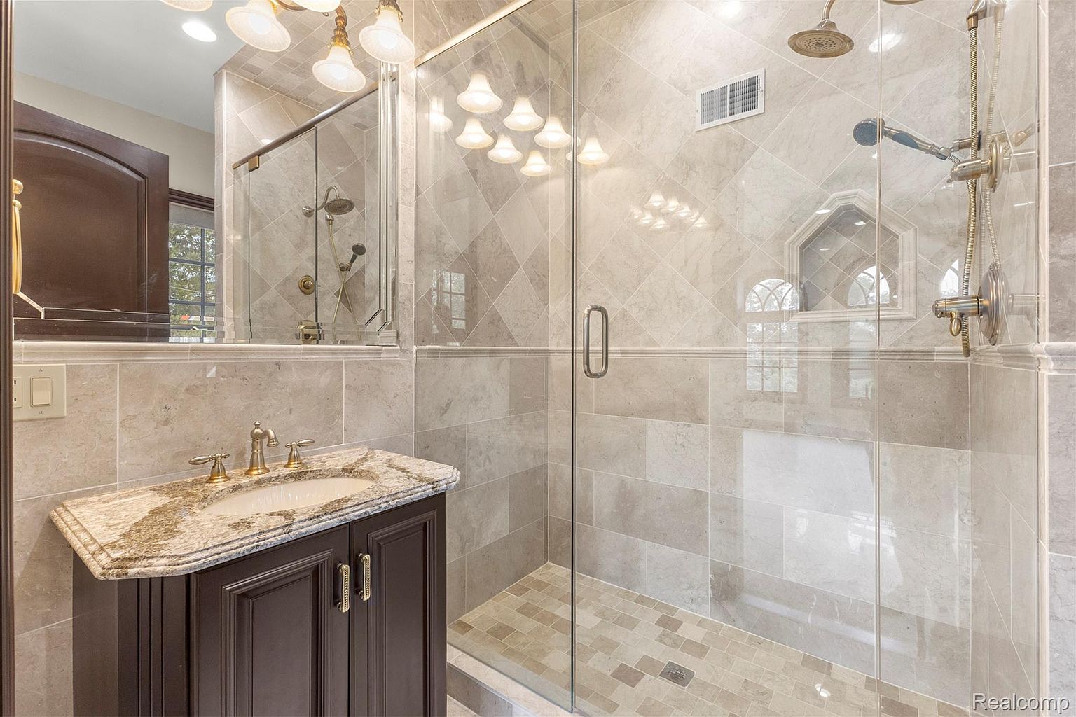 This is a luxurious primary bathroom featuring a glass-enclosed shower with dual showerheads and a built-in niche. The vanity has a dark wood cabinet with a granite countertop and gold fixtures. The walls and floor are covered in neutral-toned tiles, creating a spa-like atmosphere.
