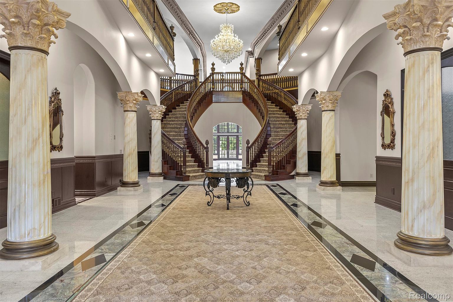 This grand foyer features a dual staircase with ornate wooden railings and carpeted steps, leading to a second-floor balcony. Marble columns with decorative capitals line the hallway, and a large chandelier hangs from the ceiling, adding to the luxurious ambiance. A patterned rug runs the length of the hallway, creating a focal point and enhancing the sense of depth.