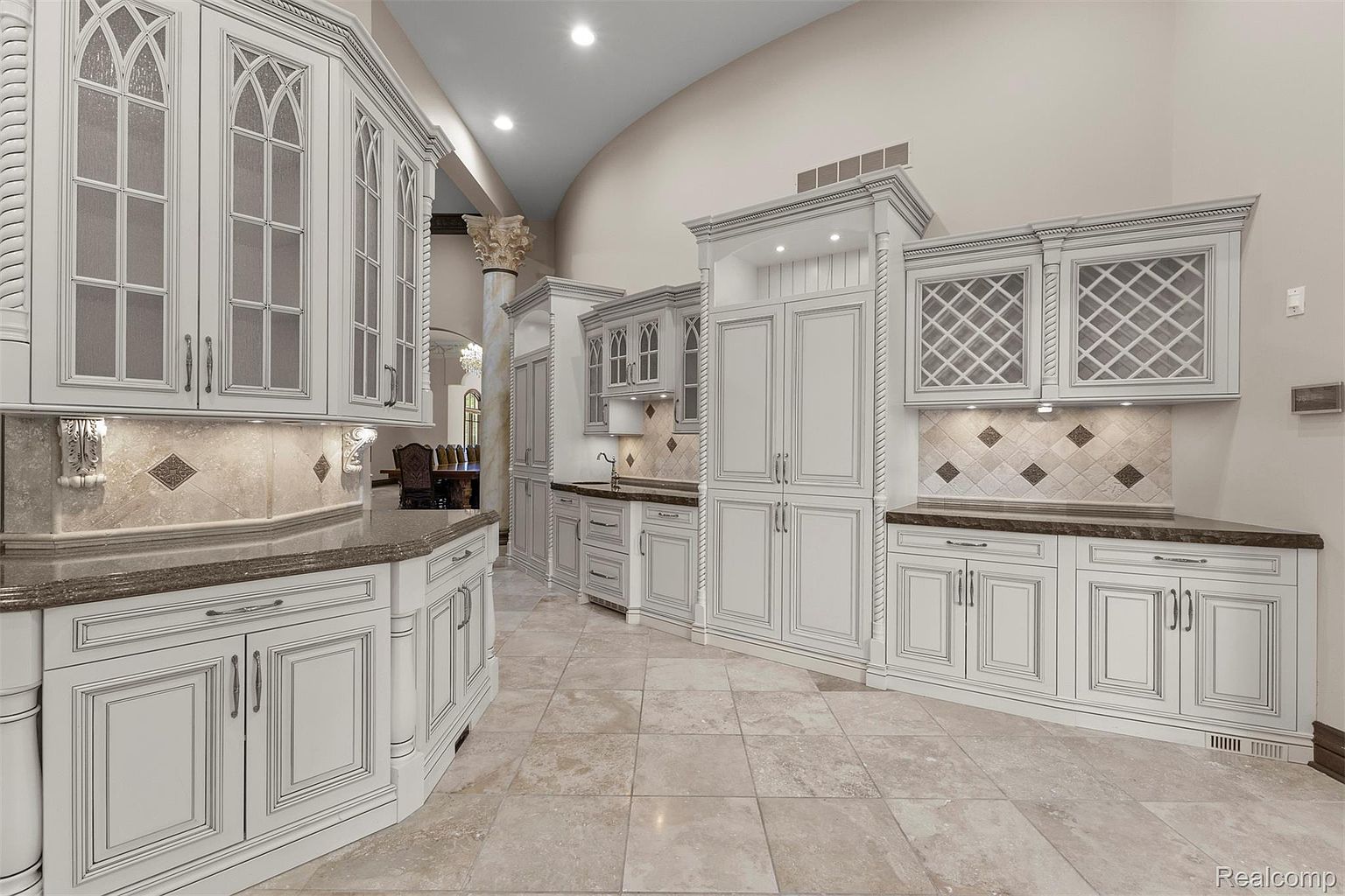 This is a spacious, luxurious kitchen featuring custom white cabinetry with detailed trim and glass-fronted upper cabinets. The countertops are a dark, contrasting material, and the backsplash features decorative tile accents. The flooring is a light-colored tile, and the overall impression is one of elegance and high-end design.