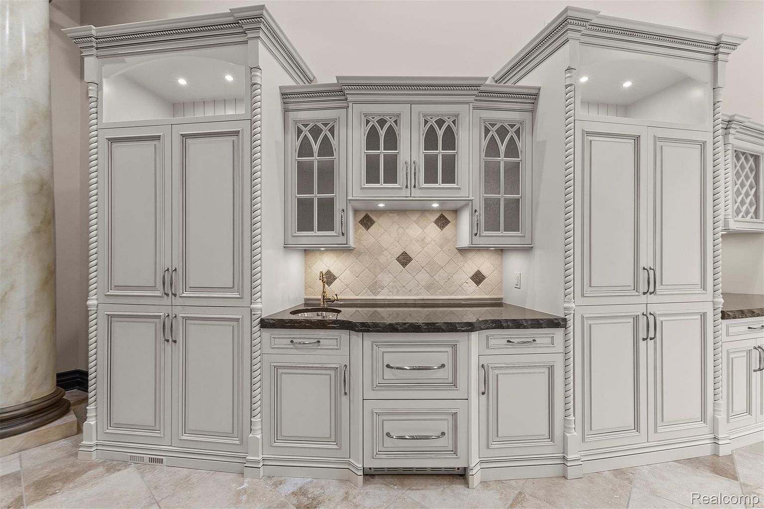 This interior shot showcases a well-appointed kitchen area with custom cabinetry in a light gray finish, complemented by dark countertops and diamond-patterned backsplash. The cabinets feature glass-fronted doors with arched detailing, and the space is illuminated by recessed lighting, creating a sophisticated and functional culinary space. The perspective is a wide shot, capturing the entire kitchen area.